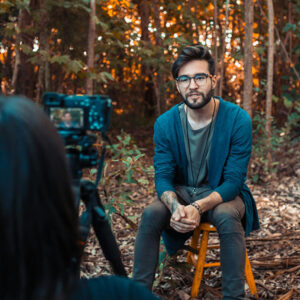 man being interviewed sitting on a stool in the autumn woods, talking into a camera